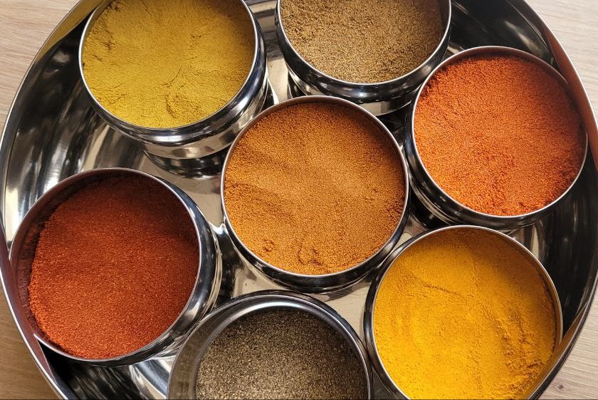 Runde Edelstahl-Schalen mit verschiedenfarbigen gemahlenen Gewürzen wie Gelb, Orange, Braun und Rot, angeordnet auf einem silbernen Tablett.